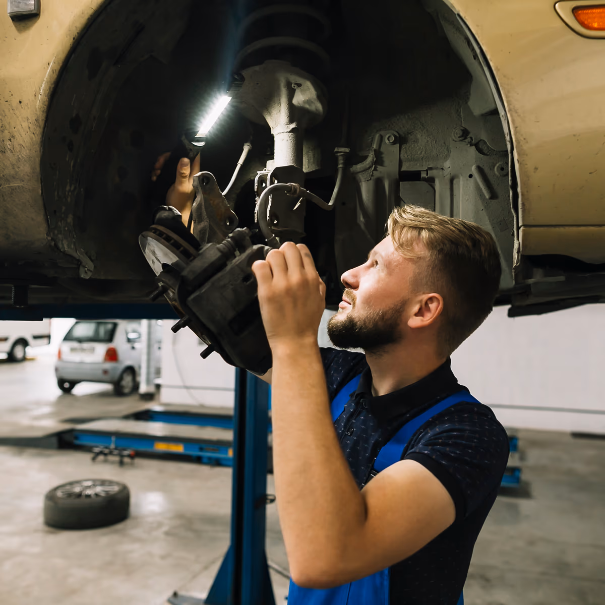 A technician working beneath a car raised on a lift, holding an inspection light up to the brake assembly