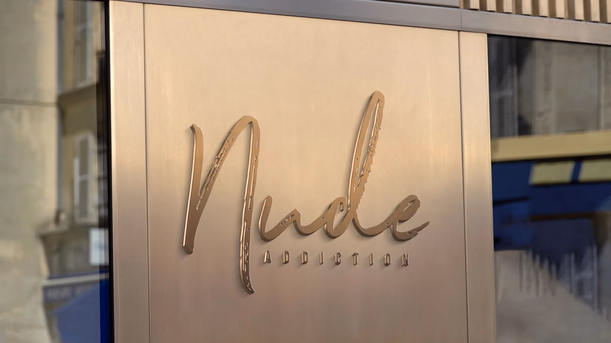 Nude Addiction embossed signage mockup showing the brand wordmark on a warm metallic facade, designed by Desque