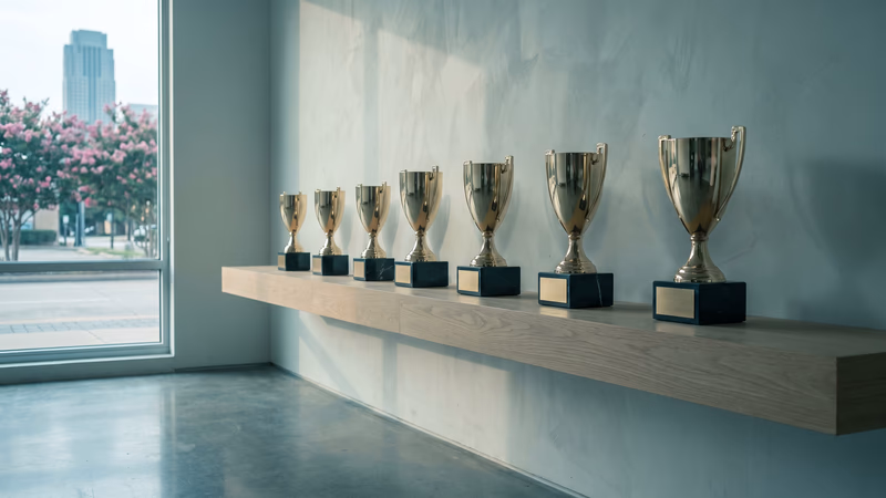 Seven gleaming gold trophies with blank, unengraved nameplates on a single wall shelf in an empty Houston room, a window beyond showing crape myrtles and a distant Uptown tower, a picture of SEO reports that look impressive but measure nothing