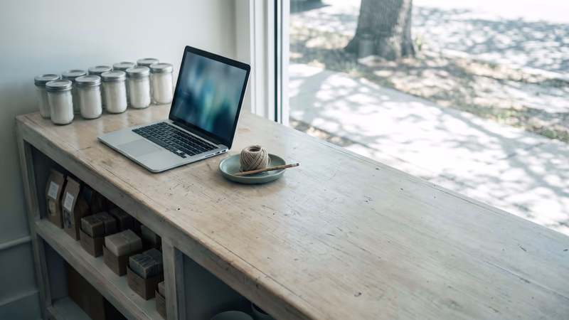 A small shop counter holding one short shelf of unlabeled products beside a single open laptop, a small catalogue run from the marketing site the business already has