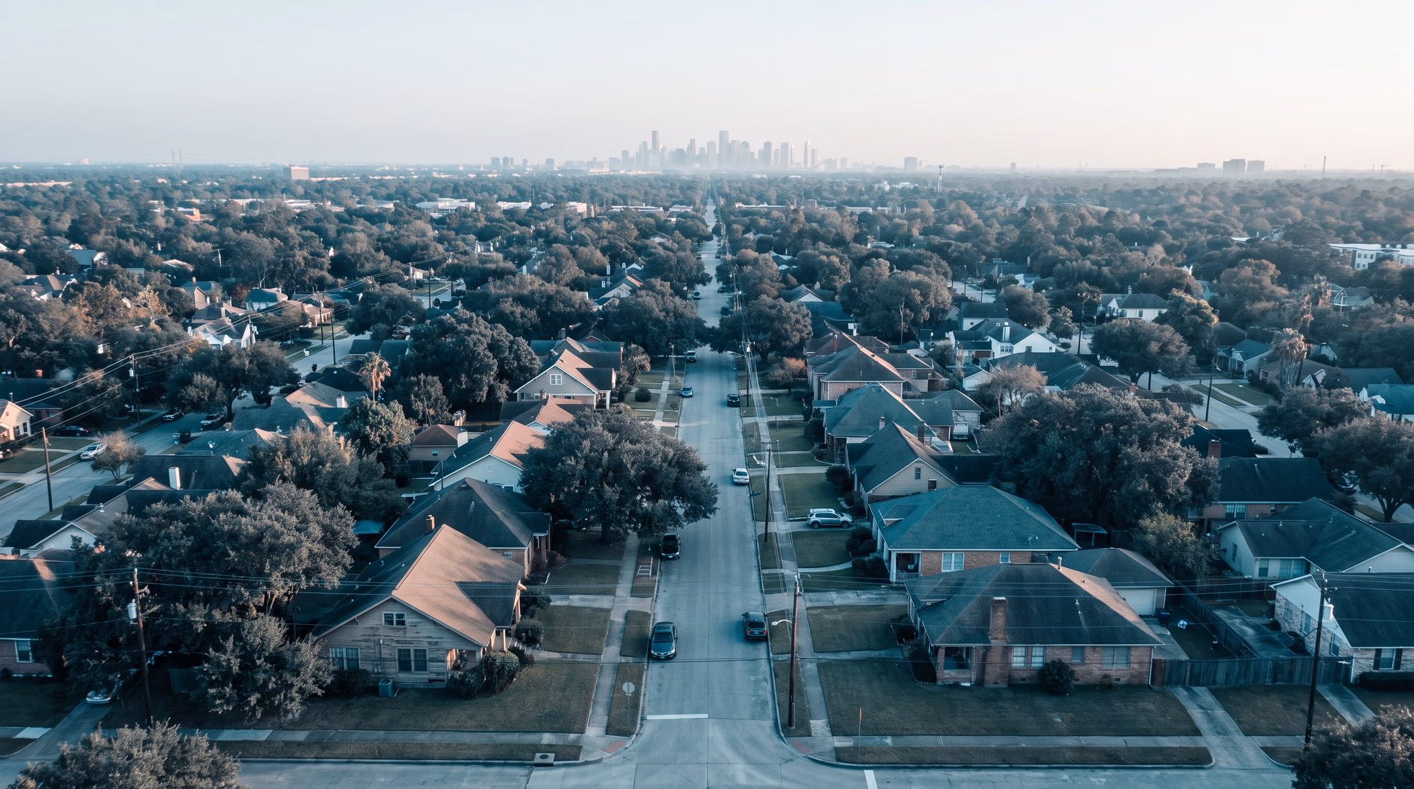 Aerial view in late-day light over a Houston neighborhood's street grid, live oaks shading the rooftops along a straight residential street that runs toward the downtown skyline hazy on the horizon