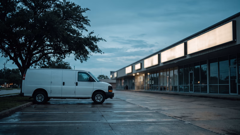 Houston strip center at blue hour after rain, a row of blank lit sign boxes above the storefronts seen edge-on, with an unmarked white service van parked under a live oak in the foreground