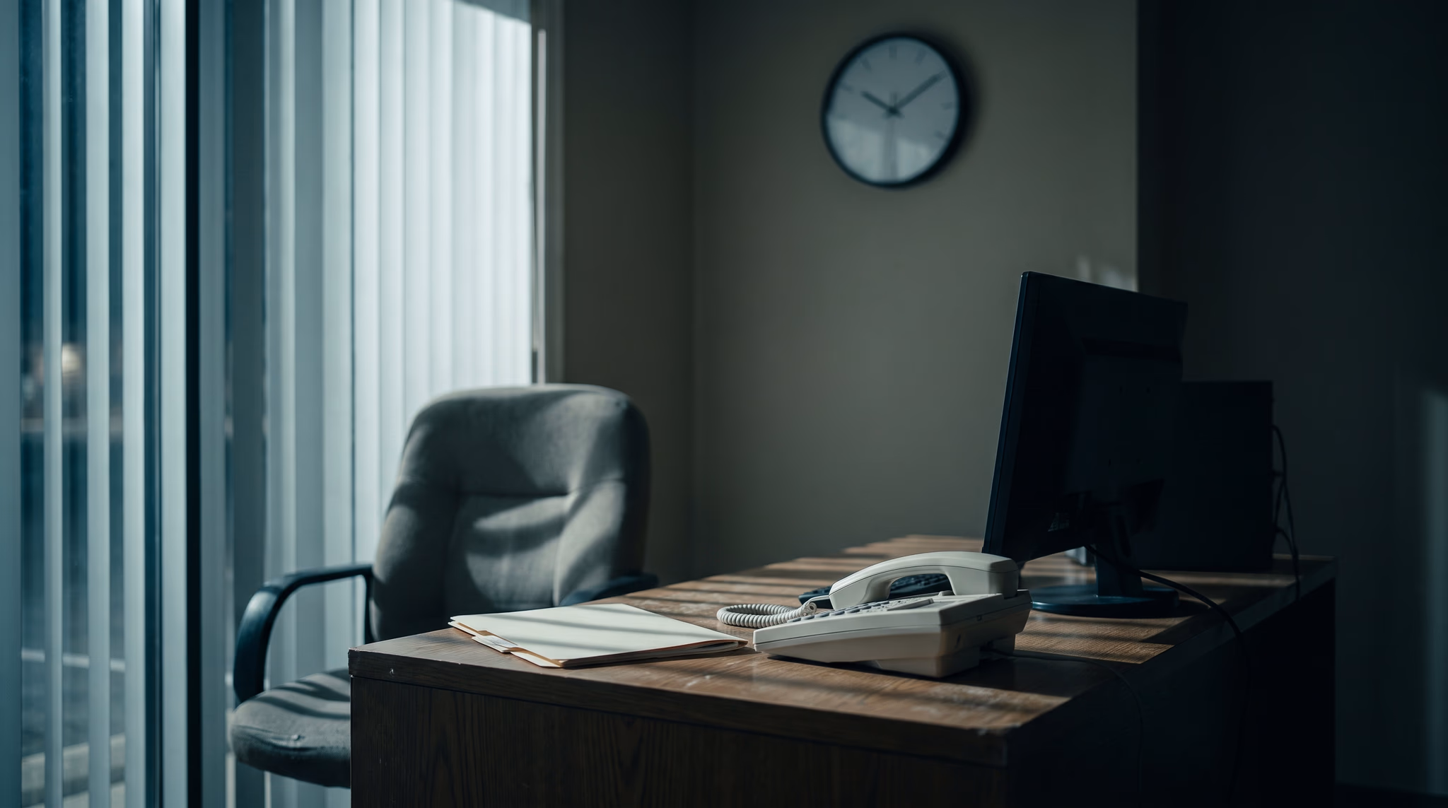 A dark small-business back office after hours, empty chair at a desk with a phone, light through half-open blinds